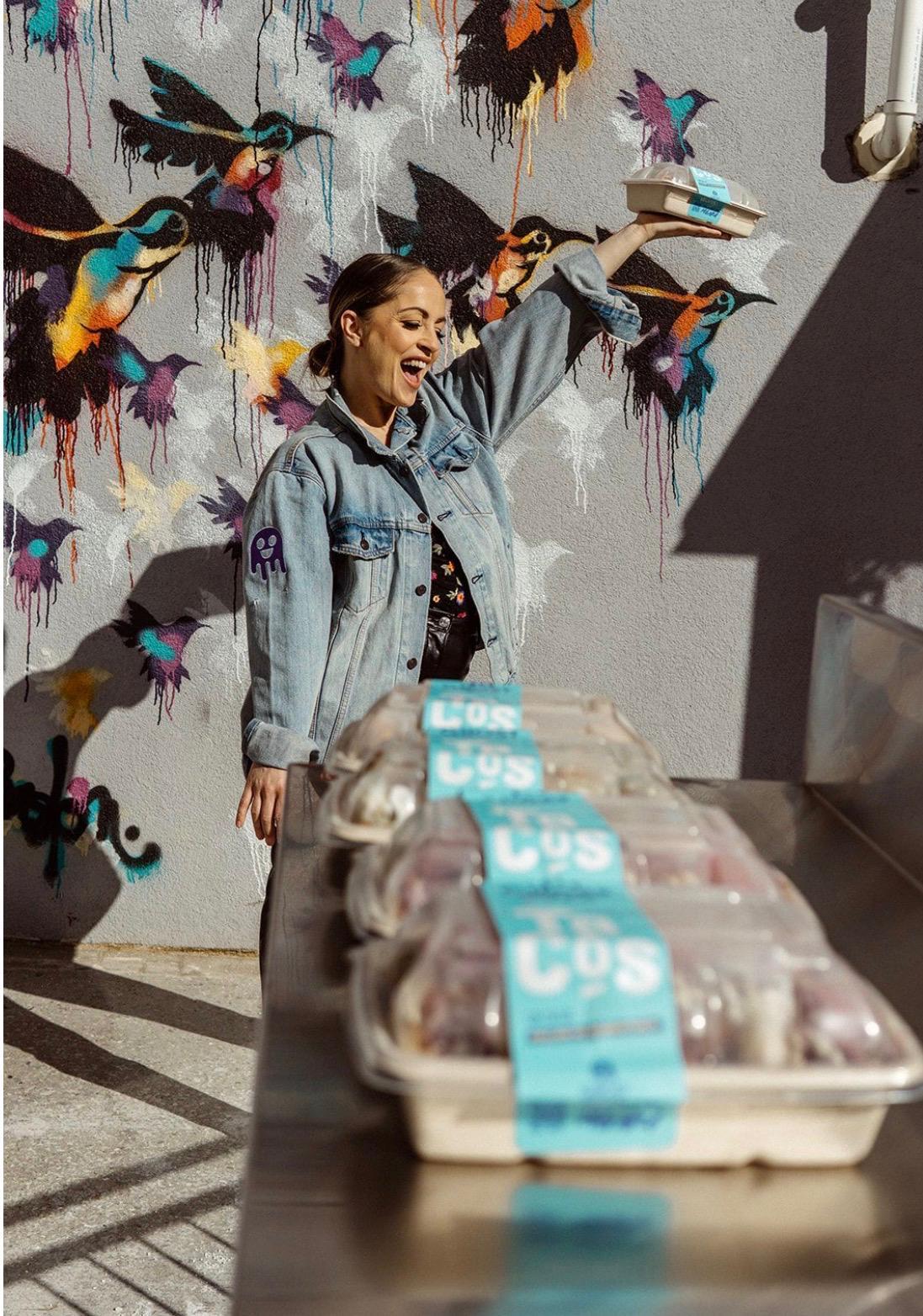 The image depicts a person standing in front of a vibrant mural, holding a box of frozen yogurt from LoS.

AI-generated content may be incorrect.